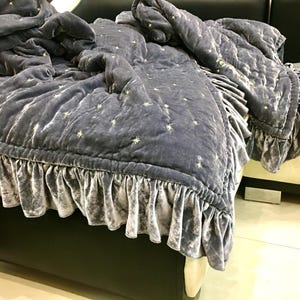 Op de afbeelding: Een stofblauwe fluwelen sprei met een gefranjerde rok. De sprei is geborduurd met kleine, goudkleurige sterren. De sprei is gedrapeerd over een donker bedframe.