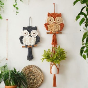 Peut inclure: Deux tentures murales en macram&eacute; en forme de hibou, l'une orange et l'autre grise, avec des accents de fil blanc. Les hiboux sont suspendus &agrave; un mur blanc avec des plantes en pots et un panier tiss&eacute; sur le sol.