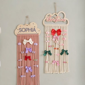 May include: Two wooden cloud-shaped organizers with the names "Sophia" and "Christina" written on them. Each organizer has a macrame hanging with multiple rows of string and various colored bows and hair clips attached.