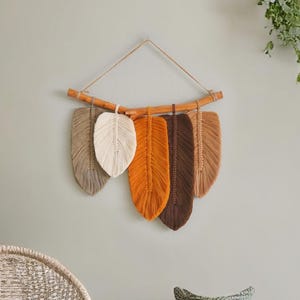 May include: A wall hanging made of five macrame feathers in shades of brown, beige, and orange. The feathers are attached to a natural branch.