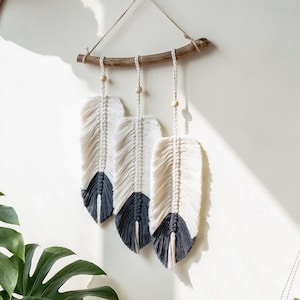 Może przedstawiać: Wisząca ozdoba ścienna z makramą z trzema białymi i szarymi piórami, wisząca na gałęzi z drewnianymi koralikami. Ozdoba ścienna jest na tle białej ściany z rośliną na pierwszym planie.
