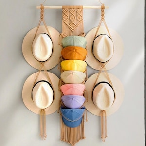 May include: A macrame wall hanging with six straw hats and seven colorful baseball caps hanging from it. The wall hanging is made of natural fiber rope and has a fringe at the bottom.