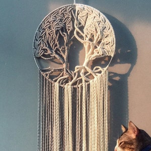 May include: A macrame wall hanging featuring a tree of life design with gray and white yarn. The hanging is in front of a gray wall with a cat sitting on a white stool.