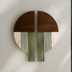 Puede incluir: Una pared moderna que cuelga hecha de madera e hilo. La madera es de color marrón oscuro y el hilo es de color verde claro y blanco. La pared que cuelga tiene la forma de un semicírculo y está dividida en dos secciones. La pared que cuelga está montada en una pared blanca.