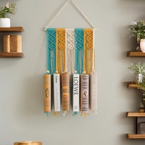 May include: A macrame wall hanging with a teal, white and mustard yellow colour scheme. The hanging has six wooden dowels that hold books in place.