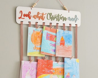 May include: A white wooden sign with the text "Look what Christina made" hanging on a wall with a pink string and wooden beads. The sign has clips to hold colorful drawings.