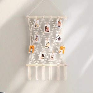 K&ouml;nnte beinhalten: Ein wei&szlig;er Makramee-Wandbehang mit einer Holzleiste und Schnurclips zum Aufh&auml;ngen von Fotos. Der Wandbehang hat ein Diamantenmuster und ist mit Quasten verziert.