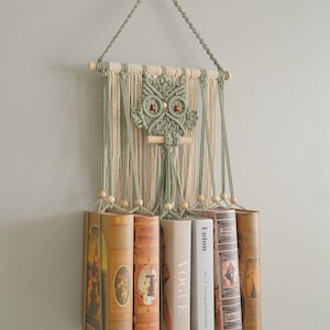 May include: A green and white macrame owl wall hanging with a wooden dowel and beads. The owl is holding a wooden dowel in its beak. The wall hanging is holding up a row of books, including a copy of Vogue magazine.