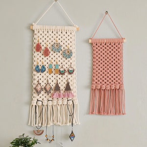 May include: Two macrame wall hangings, one white with a geometric design and the other pink with a more intricate pattern. The white hanging has a variety of earrings and necklaces hanging from it.