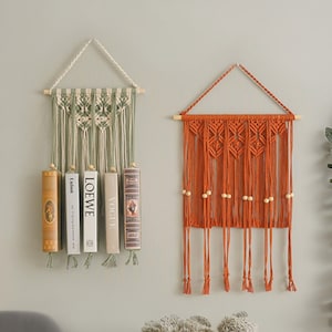 May include: Two macrame wall hangings, one in green and white with books, the other in orange with wooden beads. Both hangings have fringe details.