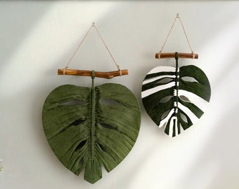 May include: Two macrame wall hangings in the shape of monstera leaves. One is green and the other is white with black stripes.