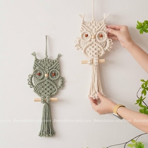 Op de afbeelding: Twee macramé wandhangers in de vorm van uilen, één in groen en één in wit, met houten latten en bruine kralenogen.