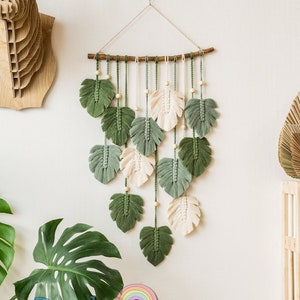 May include: A green and white macrame wall hanging with a leaf design. The wall hanging is made of yarn and is hung from a wooden dowel.