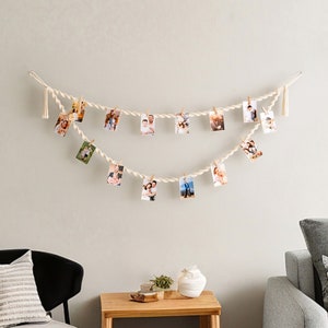 May include: A white rope photo display with tassels hangs on a white wall. The rope is strung with small photos of people, and there are wooden clothespins holding the photos in place. A gray couch with pillows sits in the foreground.