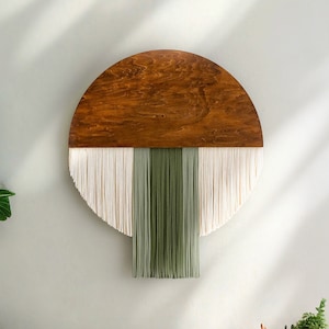 Op de afbeelding: Een houten halfronde wandhanger met een franje van wit en groen garen. Het hout is donkerbruin van kleur en de franje hangt aan de onderkant van de cirkel.