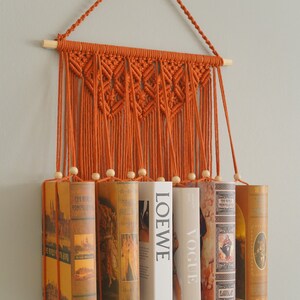 May include: A macrame wall hanging with a geometric pattern in orange. The hanging is used to hold books in place. The books are a mix of hardback and paperback and have various titles and covers.