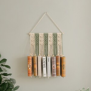 May include: A macrame wall hanging with a green and white pattern, holding a row of books. The books have titles like "LOEWE" and "Vogue".