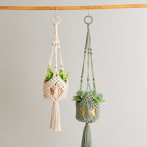 Puede incluir: Dos maceteros colgantes de macramé con listones de madera, uno es de color blanco natural y el otro es verde salvia. Ambos maceteros tienen una maceta pequeña con una planta dentro.