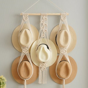 以下が含まれることがあります： マクラメ製の帽子掛けに、麦わら帽子が5つ掛けられています。帽子掛けは、天然の綿ロープで作られており、上部に木製の棒があります。帽子は、茶色とベージュの様々な色合いです。