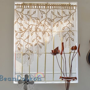 May include: A white macrame wall hanging with a leaf design hangs in a window. A black acoustic guitar hangs on the wall to the right of the window. A vase with brown lotus flowers sits on a windowsill.