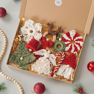 Könnte beinhalten: Eine Geschenkschachtel aus Pappe, gefüllt mit verschiedenen Makramee-Weihnachtsanhängern, darunter eine weiße Schneeflocke, ein grüner Baum, eine rote Glocke, ein braunes Rentier, ein grüner Kranz, ein rot-weiß gestreiftes Bonbon, ein Engel, eine Zuckerstange und ein Wichtel.