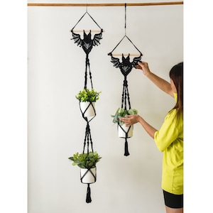 Pode incluir: Dois suportes de plantas de macramé pretos com desenhos de morcegos, cada um com um vaso branco com uma planta verde. Os suportes estão pendurados em uma haste de madeira contra uma parede branca.