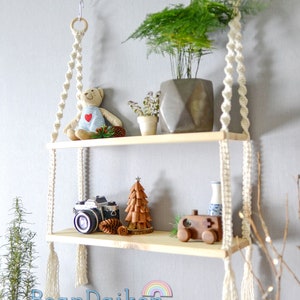 May include: Two-tiered wooden shelf with macrame hanging cords and tassels. The shelf is decorated with a teddy bear, a small potted plant, a miniature Christmas tree, a camera, pinecones, a wooden toy truck, and a small blue and white vase.