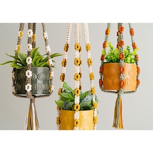 Op de afbeelding: Drie macramé plantenhangers met potten en planten in verschillende kleuren. De hangers zijn gemaakt van een beige en wit koord met witte en oranje bloemen. De potten zijn groen, oranje en geel.