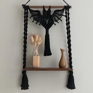 Puede incluir: Colgante de pared de macramé negro con diseño de murciélago, con una barra de madera y borlas negras. El colgante se muestra en un estante con un jarrón de flores secas y un jarrón de cerámica marrón.