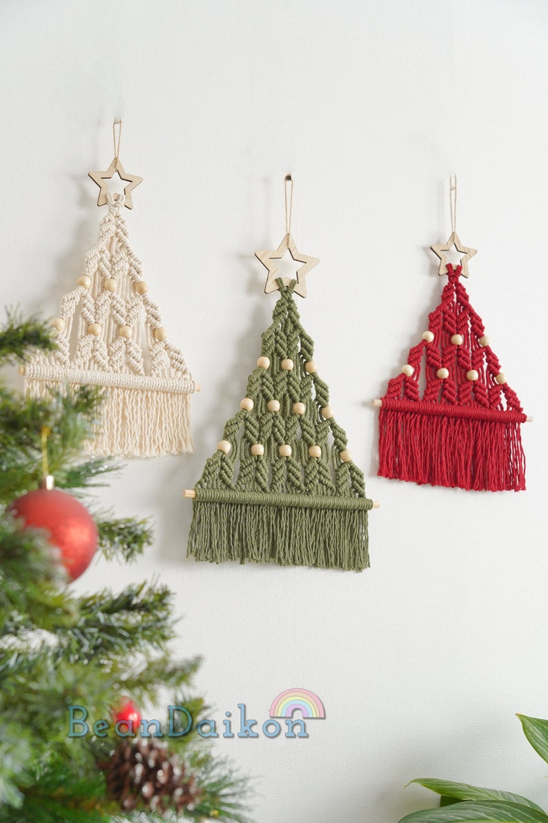 Puede incluir: Tres decoraciones de pared de &aacute;rbol de Navidad de macram&eacute; hechas a mano con cuentas de madera y adornos de estrellas. Los &aacute;rboles son de color crema, verde y rojo.