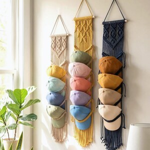 May include: Three macrame hat organizers in beige, yellow, and navy blue hang on a white wall. Each organizer holds several baseball caps in various colors, including yellow, green, pink, and blue. The organizers have a fringe detail at the bottom.