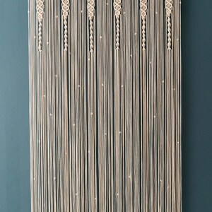 K&ouml;nnte beinhalten: Ein Wandbehang aus Makramee in beige mit einem geometrischen Muster. Der Behang hat kleine wei&szlig;e Perlen, die in das Design eingearbeitet sind.