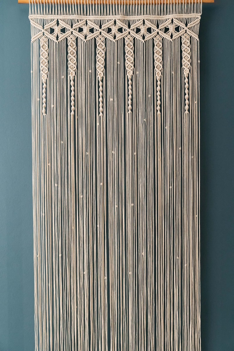K&ouml;nnte beinhalten: Ein Wandbehang aus Makramee in beige mit einem geometrischen Muster. Der Behang hat kleine wei&szlig;e Perlen, die in das Design eingearbeitet sind.