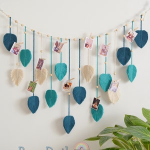 K&ouml;nnte beinhalten: Eine Wanddekoration mit einer Schnur aus teal- und cremefarbenen Garnbl&auml;ttern mit Holzperlen und W&auml;scheklammern, die Fotos halten. Die Bl&auml;tter h&auml;ngen an einer Schnur aus Holzperlen. Der Text "BeanDaikon" ist in der linken unteren Ecke des Bildes sichtbar.