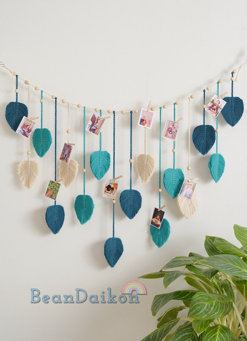 K&ouml;nnte beinhalten: Eine Wanddekoration mit einer Schnur aus teal- und cremefarbenen Garnbl&auml;ttern mit Holzperlen und W&auml;scheklammern, die Fotos halten. Die Bl&auml;tter h&auml;ngen an einer Schnur aus Holzperlen. Der Text "BeanDaikon" ist in der linken unteren Ecke des Bildes sichtbar.