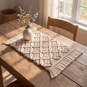 May include: A cream-colored macrame table runner with a geometric diamond pattern and fringed edges. A small white vase with dried flowers sits on the runner, displayed on a rustic wooden table. The table is near a window.