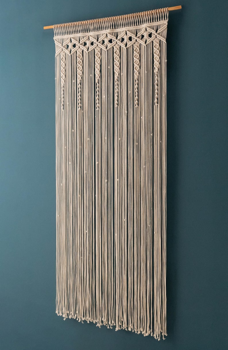 K&ouml;nnte beinhalten: Ein Makramee-Wandbehang mit geometrischem Muster und einer Holzleiste oben. Der Behang ist aus naturfarbenem Baumwollgarn gefertigt und hat kleine wei&szlig;e Perlen, die in das Muster eingearbeitet sind.