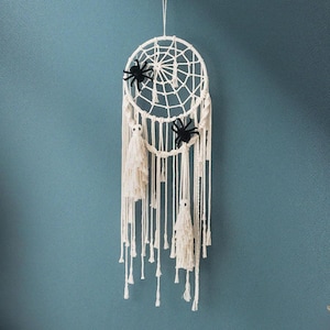 May include: A white macrame dream catcher with two black spiders and two white tassels. The dream catcher has a circular design with a web-like pattern in the center. The tassels hang down from the bottom of the dream catcher.