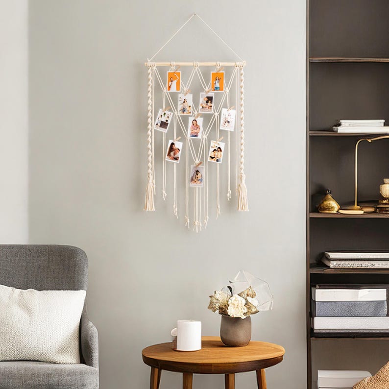 Pode incluir: Uma tape&ccedil;aria de macram&eacute; branca com uma barra de madeira e v&aacute;rios clipes para fotos. A tape&ccedil;aria de parede &eacute; decorada com fotos pequenas de pessoas e est&aacute; pendurada em uma parede cinza claro.