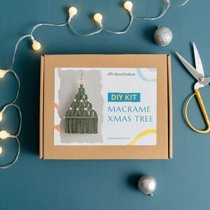 May include: A brown box with a white label that says "DIY KIT MACRAME XMAS TREE" and a picture of a green macrame Christmas tree ornament. The box is on a blue background with white lights and silver ornaments.