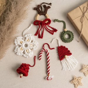 Op de afbeelding: Een set van 8 handgemaakte kerstversieringen met verschillende ontwerpen, waaronder een sneeuwvlok, een snoepstok, een rendier, een bel, een kabouter en een krans. De versieringen zijn gemaakt van wit, rood en groen garen.