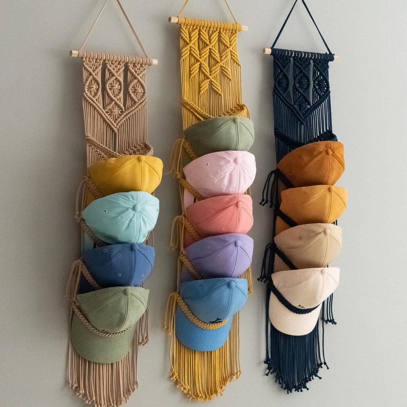 May include: Three macrame hat organizers in beige, yellow, and navy blue, each holding several baseball caps in various colors. The organizers are suspended from wooden dowels, showcasing a bohemian style and practical storage solution.