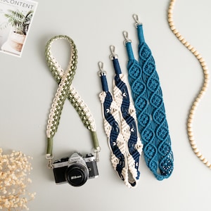 Puede incluir: Tres correas para cámara hechas de macramé con diferentes combinaciones de colores. Las correas son verde y blanco, azul y blanco, y azul. Las correas están dispuestas sobre una superficie gris claro.
