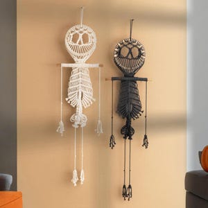 May include: Two macrame wall hangings shaped like skeletons, one in white and one in black. Each piece features a circular head with hollow eye sockets, a horizontal wooden dowel for support, and a ribcage design made of knotted cord.