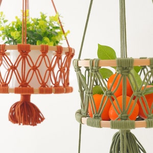 May include: Two macrame plant hangers, one orange and one green, with wooden rings. The orange hanger has a green plant inside and the green hanger has oranges inside.