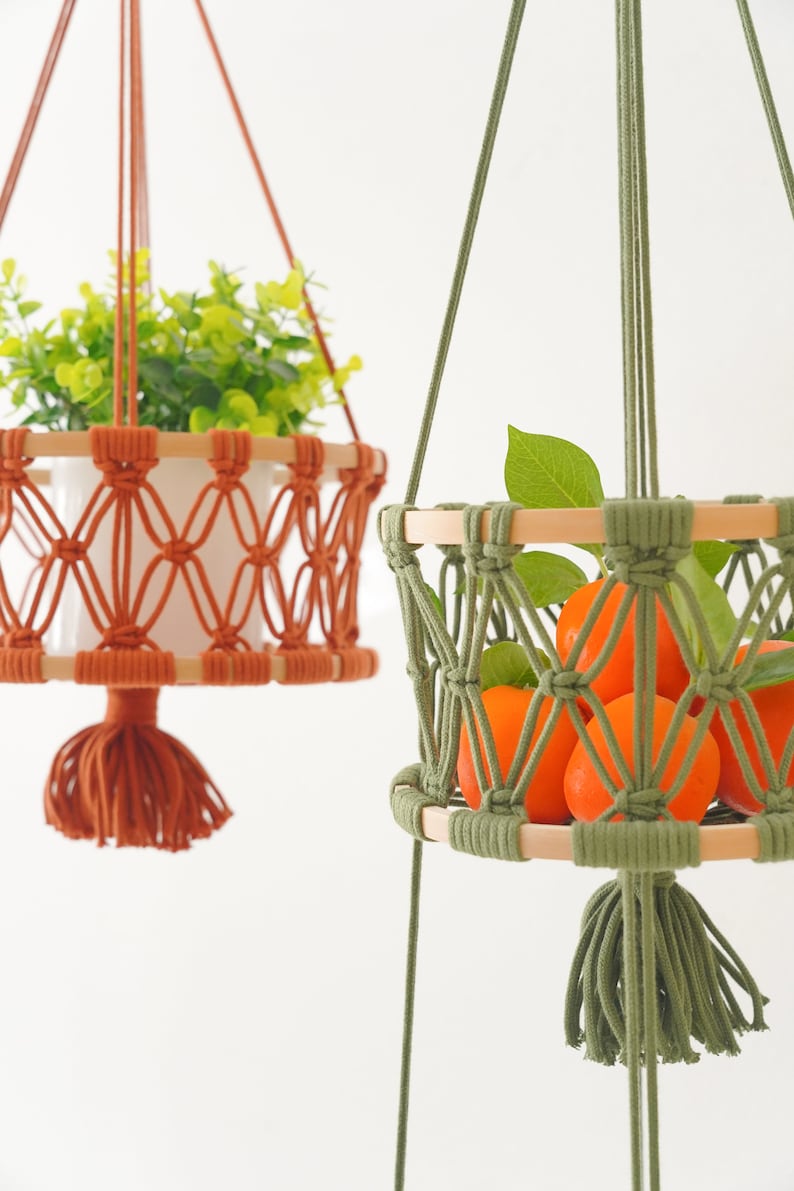 May include: Two macrame plant hangers, one orange and one green, with wooden rings. The orange hanger has a green plant inside and the green hanger has oranges inside.