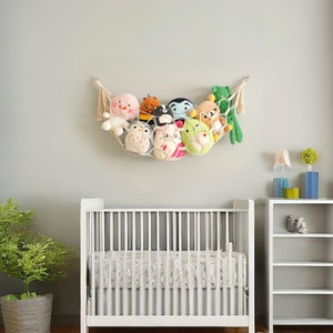 Op de afbeelding: Een wit babybedje met een wit en beige gestreept lakentje. Een witte boekenkast met drie planken staat aan de rechterkant van de afbeelding. Een witte hangmat met een verscheidenheid aan knuffels hangt aan de muur boven het babybedje.