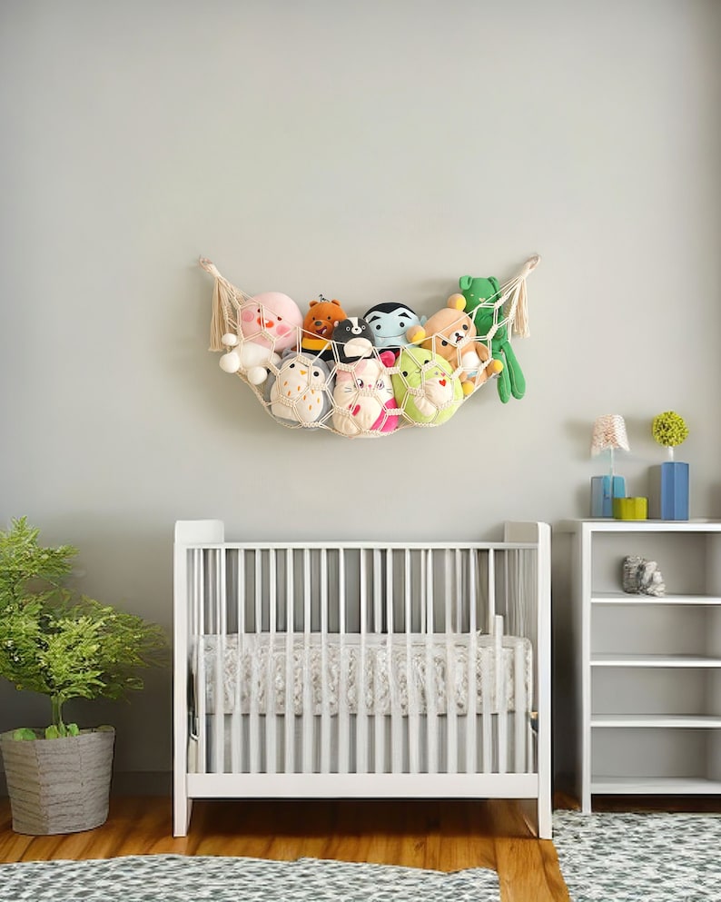 Op de afbeelding: Een wit babybedje met een wit en beige gestreept lakentje. Een witte boekenkast met drie planken staat aan de rechterkant van de afbeelding. Een witte hangmat met een verscheidenheid aan knuffels hangt aan de muur boven het babybedje.