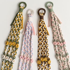 May include: Four macrame plant hangers in various colors. Each hanger features a braided design with a loop at the top and tassels at the bottom. The hangers are adorned with small, daisy-like flowers. Colors include green, yellow, white, pink, and brown.