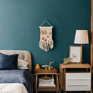 May include: A white macrame wall hanging with a geometric design and pink and white embellishments. The hanging is displayed on a teal wall above a wooden nightstand with a white lamp.